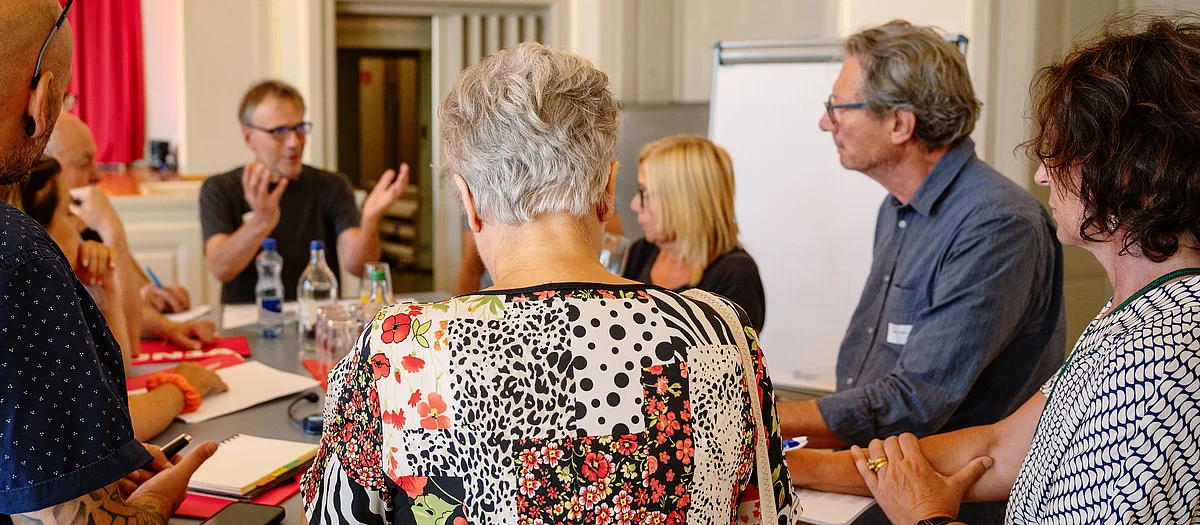 Eine Gruppe von Menschen sitzt an einem Tisch und diskutiert. Ein Mann gestikuliert, während die anderen zuhören. Im Hintergrund steht ein Flipchart.