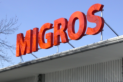 Logo de Migros en grande lettres orange sur le toit d'un bâtiment.
