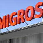 Logo de Migros en grande lettres orange sur le toit d'un bâtiment.