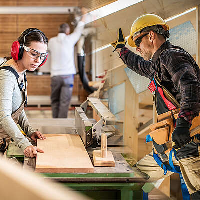Frau bedient eine Tischfräse in einer Schreinerei, daneben ein Zimmermann mit Helm und Werkzeuggurt im Holzbau.