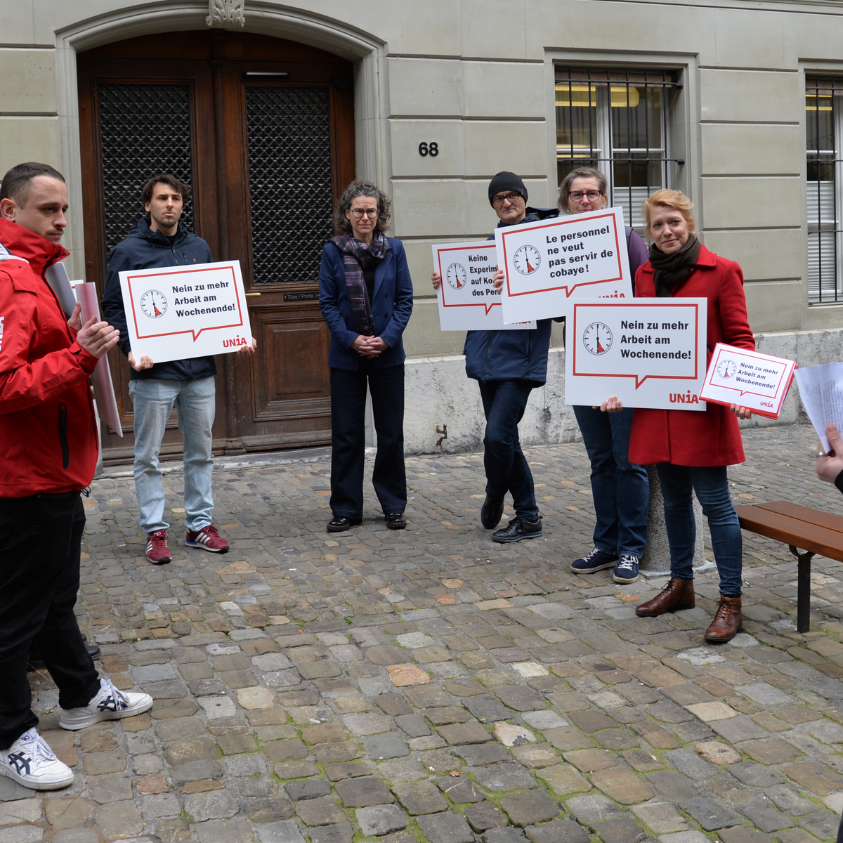 Eine Gruppe von Gewerkschafter:innen übergibt der Stadtkanzlei die Petition gegen verlängerte Ladenöffnungszeiten in der Stadt Bern. Sie halten Schilder in den Händen, auf denen «Nein zu mehr Arbeit am Wochenende» und «Keine Experimente auf Kosten des Personals» zu lesen ist. 