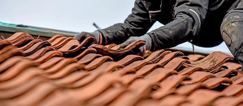 Arbeitende Person mit Handschuhen setzt eine Dachziegel auf ein Ziegeldach.