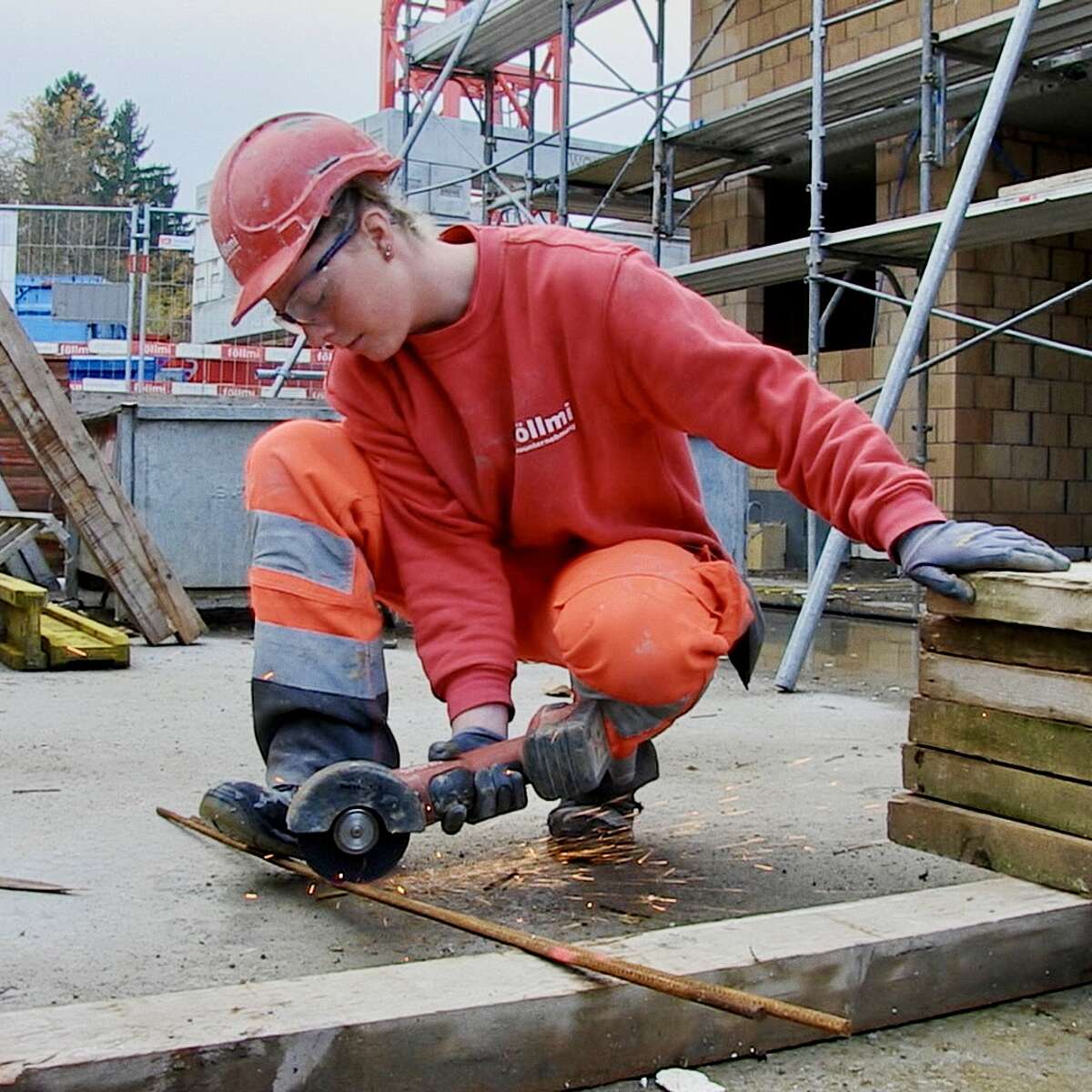 Junge Frau mit Schutzhelm, Schutzbrille und Handschuhen trennt auf einer Baustelle mit einem Winkelschleifer eine Metallstange; Funken sprühen.