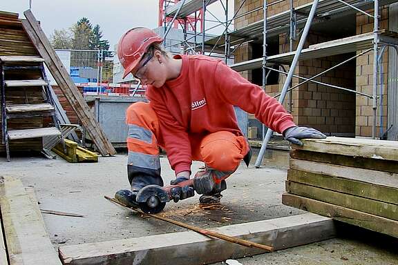 Une jeune femme portant un casque de protection, des lunettes de protection et des gants coupe une barre métallique à l'aide d'une meuleuse d'angle sur un chantier; des étincelles jaillissent.