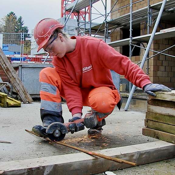 Junge Frau mit Schutzhelm, Schutzbrille und Handschuhen trennt auf einer Baustelle mit einem Winkelschleifer eine Metallstange; Funken sprühen.