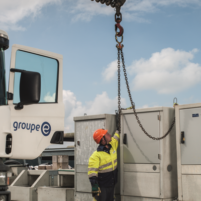 Un ouvrier avec un casque fixe une chaîne à un crochet de grue à côté d’un véhicule de service de Groupe E.