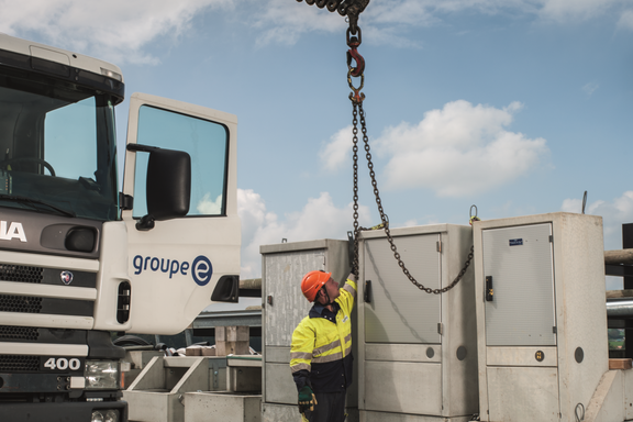 Un ouvrier avec un casque fixe une chaîne à un crochet de grue à côté d’un véhicule de service de Groupe E.