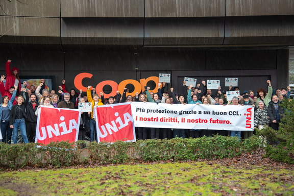 Un gruppo di 50 persone innalza uno striscione, bandiere, cartelli di cartone e grandi lettere ritagliate che riproducono il logo aziendale di Coop. Alzano il pugno al cielo e trasmettono un’immagine sindacale e combattiva. Lo slogan dello striscione è: «Più protezione grazie a un CCL forte: il nostro futuro, i nostri diritti»