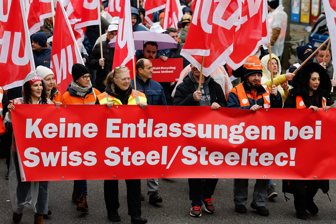 Menschen demonstrieren mit Unia-Fahnen und einem Transparen mit der Aufschrift: «Keine Entlassungen bei Swiss Steel/Steeltec!». Sie tragen Arbeitskleidung und Helme.