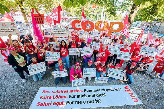 Un groupe de personnes manifeste avec des pancartes et banderoles pour des salaires équitables chez Coop. Beaucoup portent des t-shirts et casquettes rouges Unia. Le logo Coop est visible.
