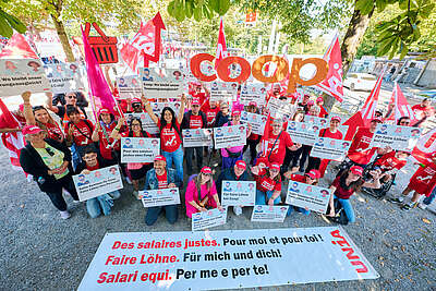 Un groupe de personnes manifeste avec des pancartes et banderoles pour des salaires équitables chez Coop. Beaucoup portent des t-shirts et casquettes rouges Unia. Le logo Coop est visible.