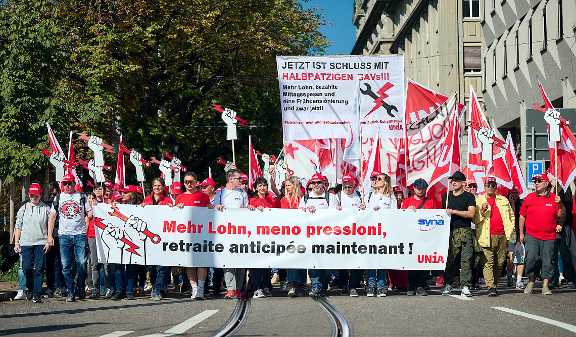 Un groupe de personnes marche avec des drapeaux et des banderoles rouges de l'Unia. La banderole de devant demande «Mehr Lohn, meno pressioni, retraite anticipée maintenant!».