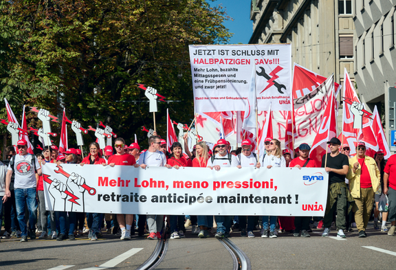 Un groupe de personnes marche avec des drapeaux et des banderoles rouges de l'Unia. La banderole de devant demande «Mehr Lohn, meno pressioni, retraite anticipée maintenant!».