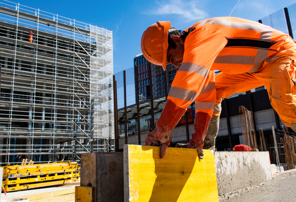 Un travailleur de la construction en tenue de protection orange soulève une plaque de bois jaune. En arrière-plan, on aperçoit un grand échafaudage.