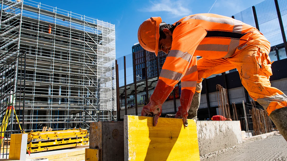 Un travailleur de la construction en tenue de protection orange soulève une plaque de bois jaune. En arrière-plan, on aperçoit un grand échafaudage.