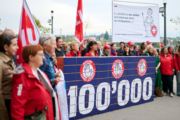 Avec d'autres organisations, Unia remet à la Chancellerie fédérale 100 000 signatures contre l'attaque sur les salaires minimums, contenues dans plusieurs cartons constituant un mur sur lequel figure le nombre 100 000.
