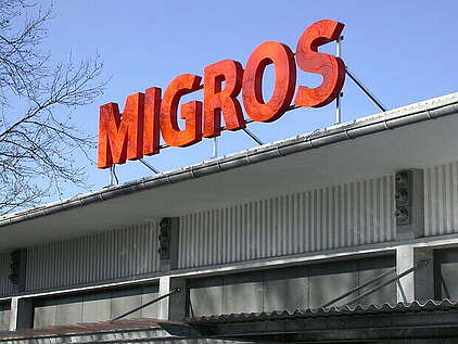 Das orange Migros-Logo auf dem Dach eines Industriegebäudes.