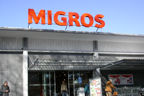 Entrée d'un magasin Migros avec des lettres lumineuses orange « MIGROS » sur fond de ciel bleu