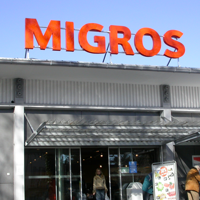 Entrée d'un magasin Migros avec des lettres lumineuses orange « MIGROS » sur fond de ciel bleu
