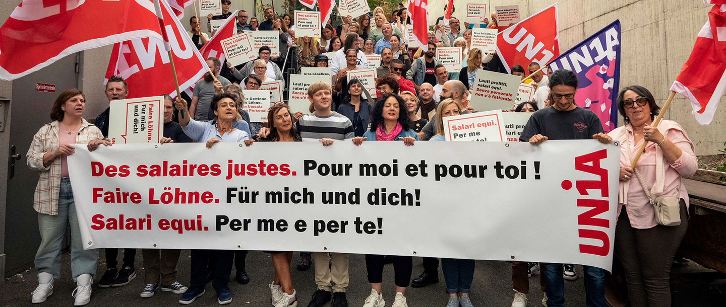 Environ 100 membres d’Unia tiennent une banderole, des pancartes et agitent des drapeaux. Sur la banderole, on peut lire: «Des salaires justes. Pour toi et pour moi!»