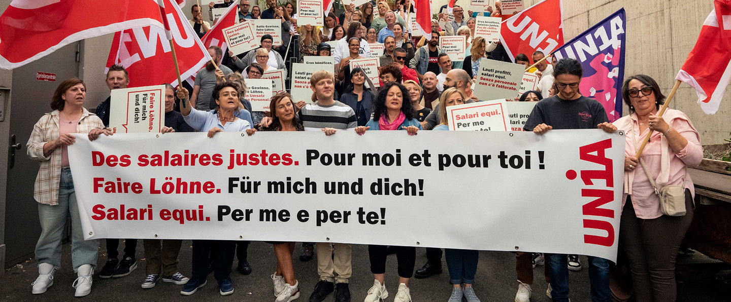Circa 100 associate e associati di Unia mostrano uno striscione e dei manifesti e sventolano delle bandiere. Lo slogan dello striscione è: «Salari equi. Per me e per te».