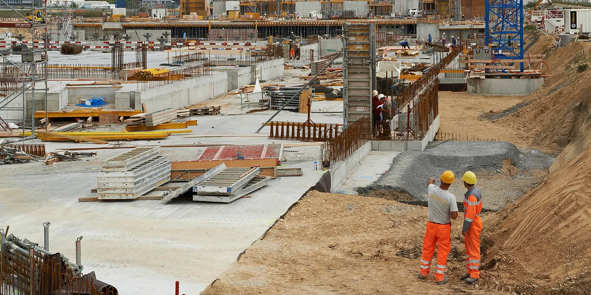 Deux contremaîtres portant des vêtements de travail orange et des casques discutent sur un grand chantier de l'avancement des travaux.  À l'arrière-plan: des grues de chantier, des fondations en béton et des matériaux de construction.