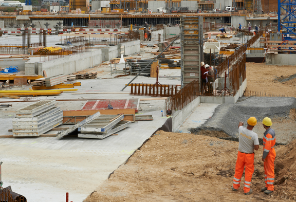 Deux contremaîtres portant des vêtements de travail orange et des casques discutent sur un grand chantier de l'avancement des travaux.  À l'arrière-plan: des grues de chantier, des fondations en béton et des matériaux de construction.