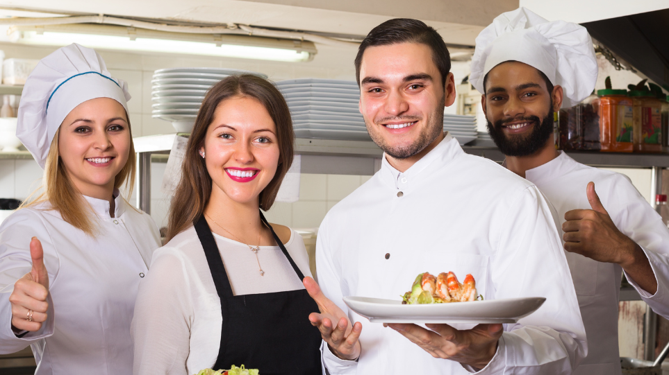 Une cuisinière, deux cuisiniers et une serveuse posent dans une cuisine de restaurant et sourient à la caméra. Deux tiennent des assiettes avec des plats, les autres montrent un pouce en l'air.