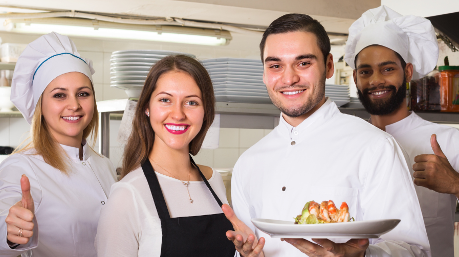 Una cuoca, due cuochi e una cameriera posano in una cucina di ristorante e sorridono alla fotocamera. Due tengono piatti con cibi, gli altri mostrano un pollice in su.