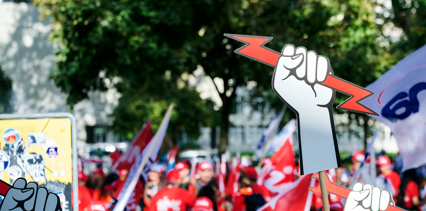 Salari più alti grazie all’impegno combattivo di elettricisti ed elettriciste - Manifestazione di elettricisti ed elettriciste; al centro: un pugno bianco in cartone tiene una saetta rossa (elettrica).