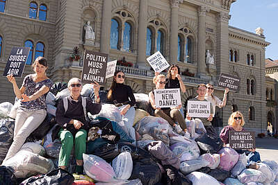 Des personnes se tiennent autour d'une montagne de vêtements devant le Parlement fédéral. Elles tiennent des pancartes et des drapeaux. Elles protestent ainsi contre l'industrie de la fast fashion, nuisible à l'environnement et à l’humain.