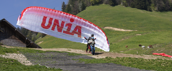 L'image montre un parapentiste se préparant au décollage. La voile du parapente est gonflée et présente un logo Unia bien visible.