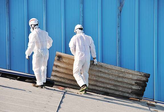 Deux travailleurs en combinaison de protection et portant des casques retirent une plaque d’amiante d’un toit.