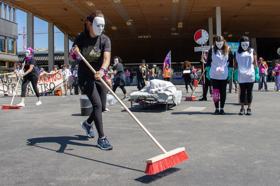 Attiviste di Unia si esibiscono in teatro di strada nell'ambito dello sciopero delle donne del 2023. Indossando maschere bianche e abiti professionali, rappresentano la pressione e lo stress a cui sono esposte nel loro lavoro di addette alle pulizie degli alberghi. La scena è ambientata davanti alla stazione ferroviaria principale di Zurigo, in Europaallee.