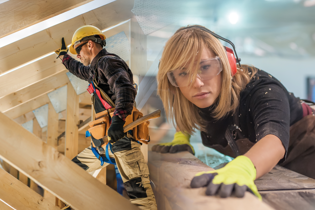 Zimmermann und Schreinerin arbeiten mit Schutzbrille, Gehörschutz und Werkzeug an Holzbalken in der Werkstatt bzw. auf der Baustelle.