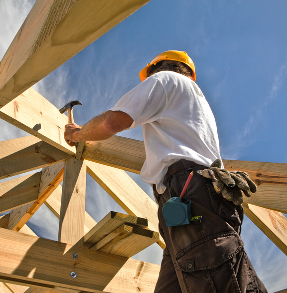 Ein Holzbauer aus der Froschperspektive fotografiert. Er hämmer an einem Holzgerüst, über ihm ein blauer Himmel mit Zirruswolken. 