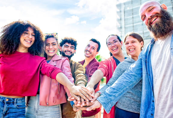 Un groupe diversifié de sept personnes debout à l'extérieur. Ils sourient et mettent leurs mains l'une sur l'autre au centre, ce qui symbolise la solidarité.