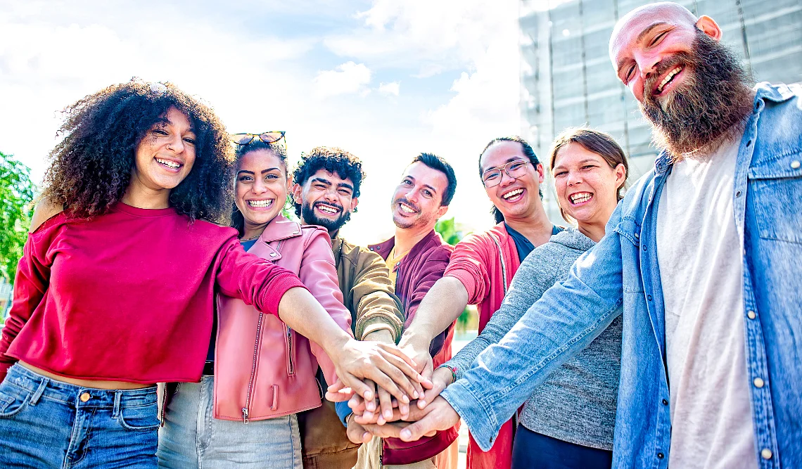 Un gruppo diversificato di sette persone che stanno all'aperto. Sorridono e mettono le loro mani una sopra l'altra al centro, il che simboleggia la solidarietà.