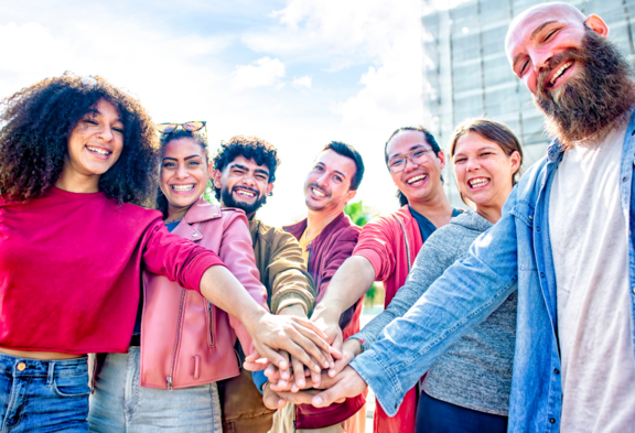 Eine diverse Gruppe von sieben Menschen, die im Freien steht.Sie lächeln und legen ihre Hände übereinander in die Mitte, was Zusammenhalt symbolisiert.