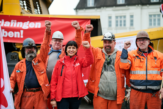 Quatre ouvriers de Stahl Gerlafingen, casqués et vêtus d'une tenue de protection orange, avec la présidente d'Unia Vania Alleva au milieu d'eux. Tous lèvent le poing de manière combative, mais avec un visage joyeux.