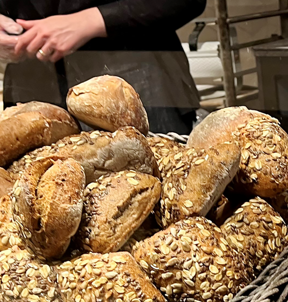 Gros plan sur des paniers en osier remplis de pains artisanaux variés, petits pains aux céréales et bretzels, avec une personne en arrière-plan portant des gants de protection dans une boulangerie.