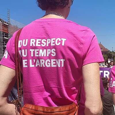 Deux personnes se tiennent à une manifestation en plein air et portent des t‑shirts roses avec les slogans «Du respect du temps et de l’argent» et «Respekt, mehr Lohn, mehr Zeit».