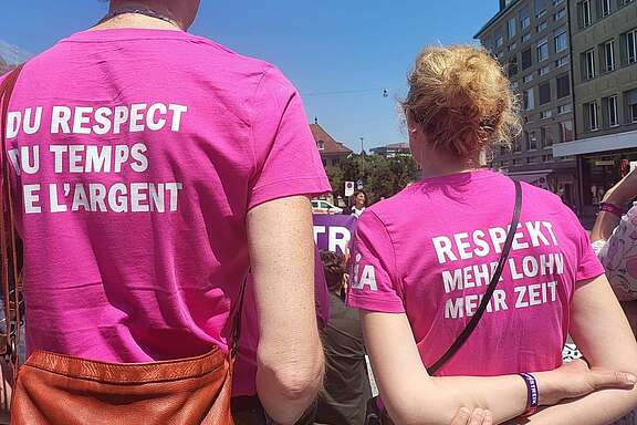 Zwei Personen stehen bei einer Demo im Freien und tragen pinkfarbene T‑Shirts mit den Aufschriften „Du respect du temps et l’argent“ und „Respekt, mehr Lohn, mehr Zeit“.