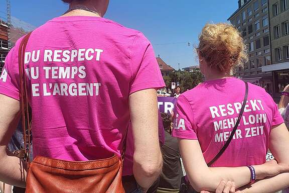 Deux personnes se tiennent à une manifestation en plein air et portent des t‑shirts roses avec les slogans «Du respect du temps et de l’argent» et «Respekt, mehr Lohn, mehr Zeit».