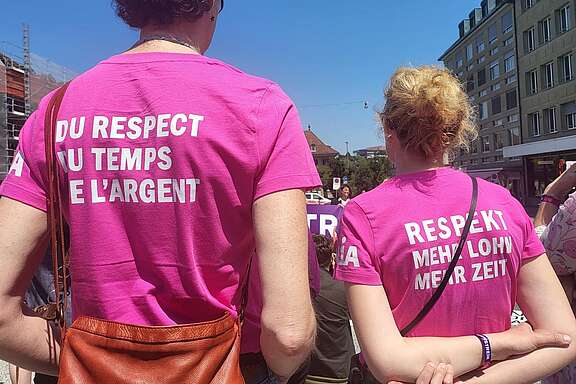 Zwei Personen stehen bei einer Demo im Freien und tragen pinkfarbene T‑Shirts mit den Aufschriften „Du respect du temps et l’argent“ und „Respekt, mehr Lohn, mehr Zeit“.