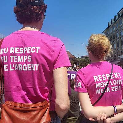 Zwei Personen stehen bei einer Demo im Freien und tragen pinkfarbene T‑Shirts mit den Aufschriften „Du respect du temps et l’argent“ und „Respekt, mehr Lohn, mehr Zeit“.
