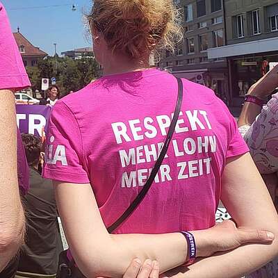 Zwei Personen stehen bei einer Demo im Freien und tragen pinkfarbene T‑Shirts mit den Aufschriften „Du respect du temps et l’argent“ und „Respekt, mehr Lohn, mehr Zeit“.