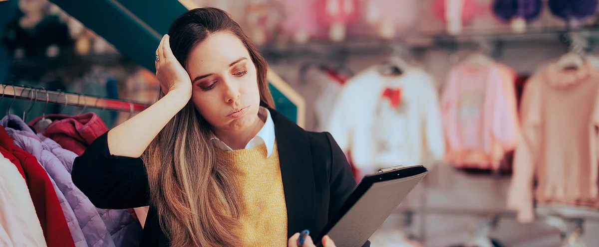Eine erschöpfte Verkäuferin mit einem Tablet in der Hand steht vor einem Kleiderständer und hält sich den Kopf - ein Zeichen von Stress wegen langer Ladenöffnungszeiten im Detailhandel.