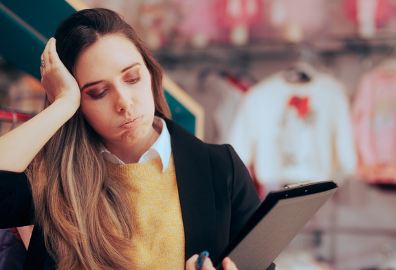 Une vendeuse épuisée, sa tablette à la main, debout devant un support à vêtements, se tient la tête: un signe de stress dû aux longues heures d’ouverture des magasins dans le commerce de détail.
