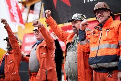 Vier Arbeiter der Stahl-Industrie in oranger Schutzkleidung erheben die Fäuste. Im Hintergrund ist eine rot-weisse Unia-Fahne sichtbar, an einer grossen Industriemaschine.